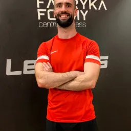 Uśmiechnięty mężczyzna w czerwonej koszulce sportowej z białymi paskami na rękawach i czarnych spodenkach, stoi na tle ściany z logo 'Fabryka Formy' i częściowo widocznym napisem 'LE'.