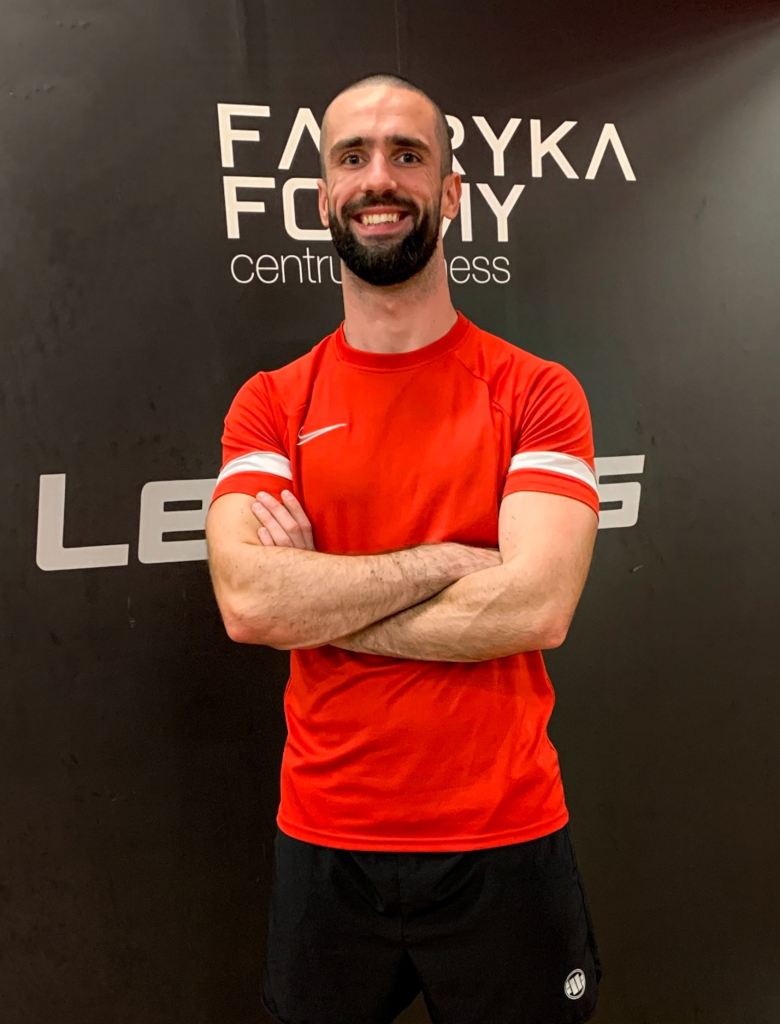 Uśmiechnięty mężczyzna w czerwonej koszulce sportowej z białymi paskami na rękawach i czarnych spodenkach, stoi na tle ściany z logo 'Fabryka Formy' i częściowo widocznym napisem 'LE'.