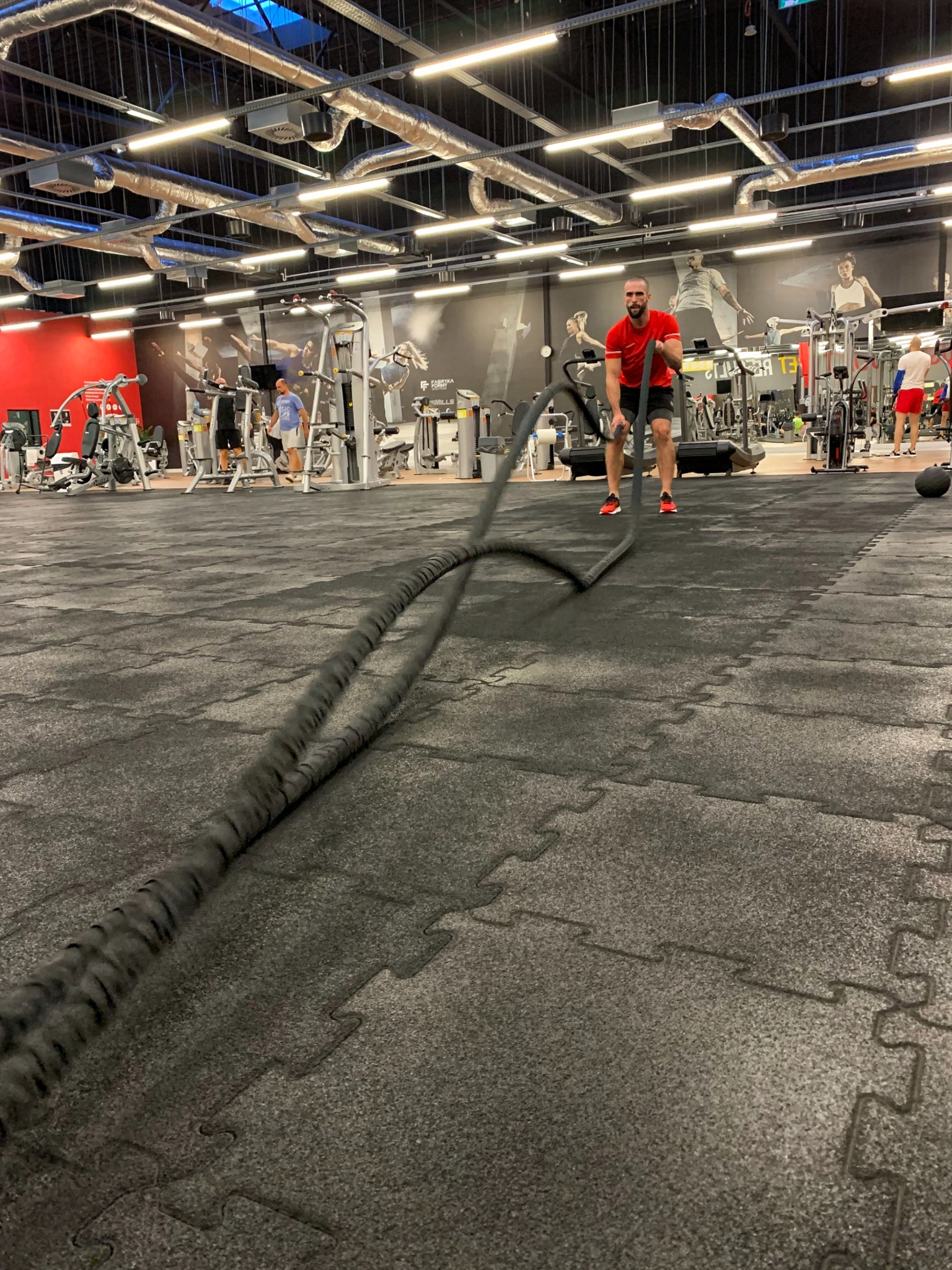 Mężczyzna w czerwonej koszulce ćwiczy z grubymi linami w nowoczesnej siłowni z widocznymi urządzeniami do ćwiczeń i innymi osobami trenującymi w tle.