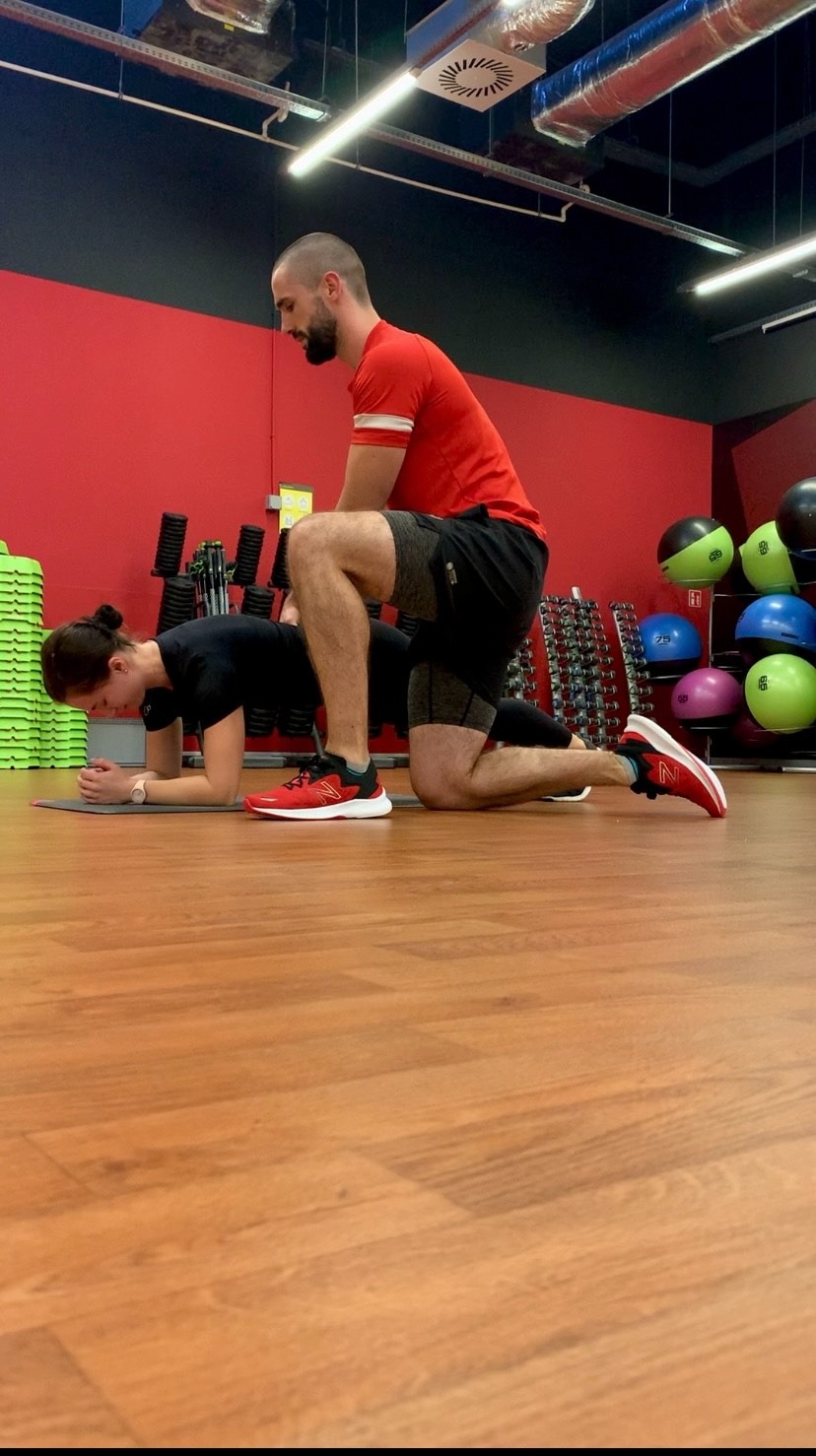 Kobieta w pozycji plank na macie, mężczyzna w czerwonej koszulce na kolanach za nią, prawdopodobnie korygujący jej postawę, siłownia w tle.