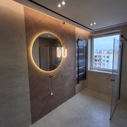 DOMIRAJ USŁUGI REMONTOWO BUDOWLANE SŁAWOMIR MARAJ - Nowoczesna łazienka z okrągłym lustrem podświetlanym LED, geometryczną ścianą z płytek, czarnym grzejnikiem drabinkowym i kabiną prysznicową ze złotą armaturą.