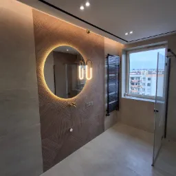 Nowoczesna łazienka z okrągłym lustrem podświetlanym LED, geometryczną ścianą z płytek, czarnym grzejnikiem drabinkowym i kabiną prysznicową ze złotą armaturą.