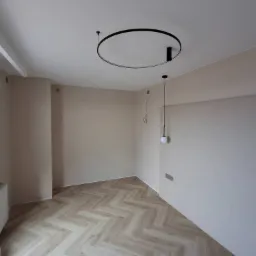 Jasne, puste pomieszczenie po remoncie z podłogą ułożoną w jodełkę, ścianami w odcieniach beżu, nowoczesnym okrągłym oświetleniem sufitowym i grzejnikiem przy oknie z butelką wody mineralnej obok.