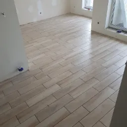 Ułożona podłoga z płytek imitujących drewno w jasnym odcieniu, widoczne ślady po gładzi na ścianach i niezabezpieczone otwory na gniazdka.