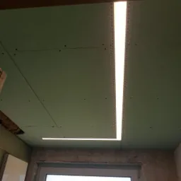 Sufit z płyt kartonowo-gipsowych z zamontowanym oświetleniem LED w profilach, widoczne ślady szpachlowania i okno w tle.