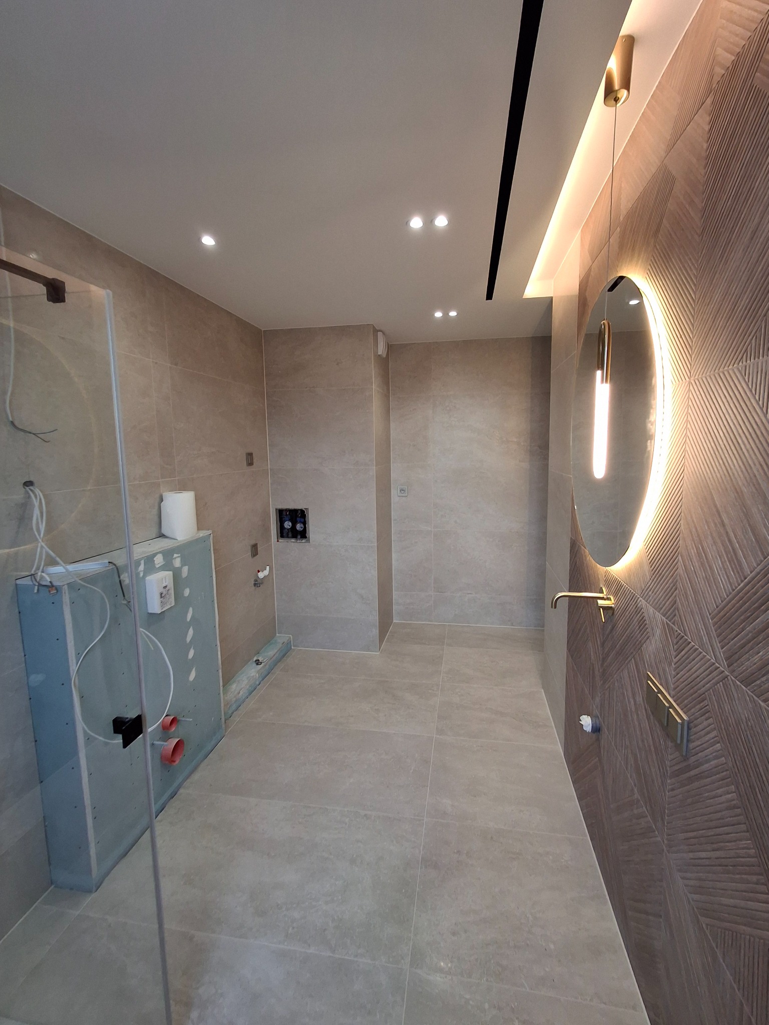 Łazienka w trakcie prac wykończeniowych: widoczna zabudowa stelaża WC z płyt karton-gips, częściowo położone płytki na ścianach i podłodze, oraz designerskie lustro z podświetleniem LED i armatura...