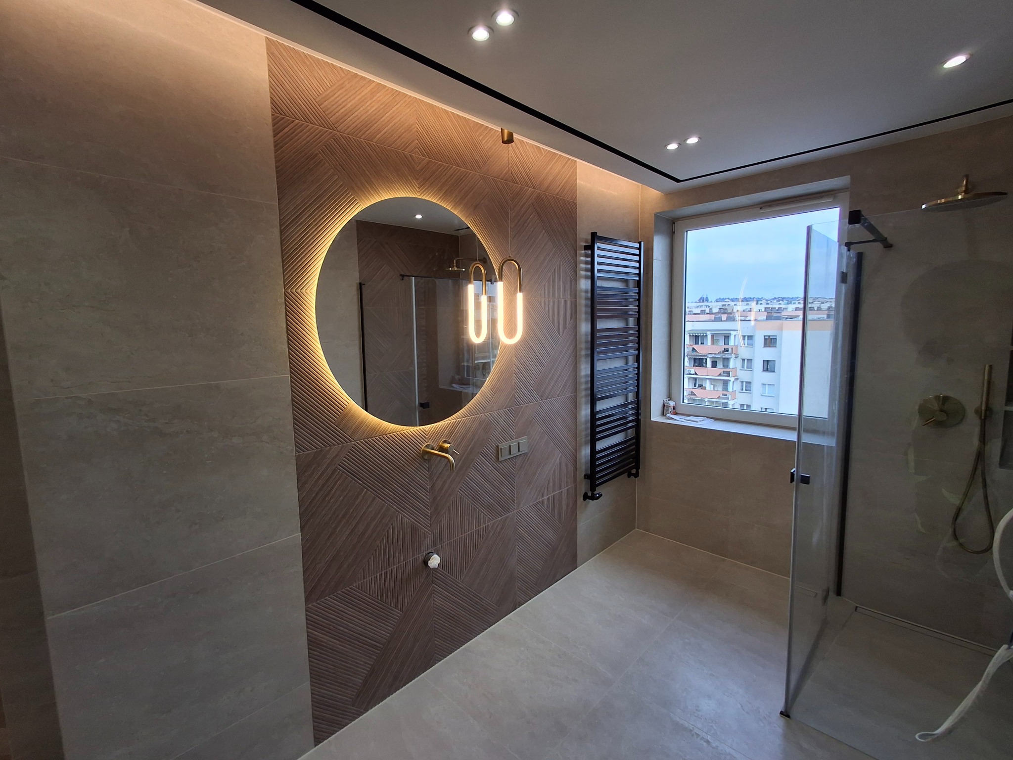 Nowoczesna łazienka z okrągłym lustrem podświetlanym LED, geometryczną ścianą z płytek, czarnym grzejnikiem drabinkowym i kabiną prysznicową ze złotą armaturą.