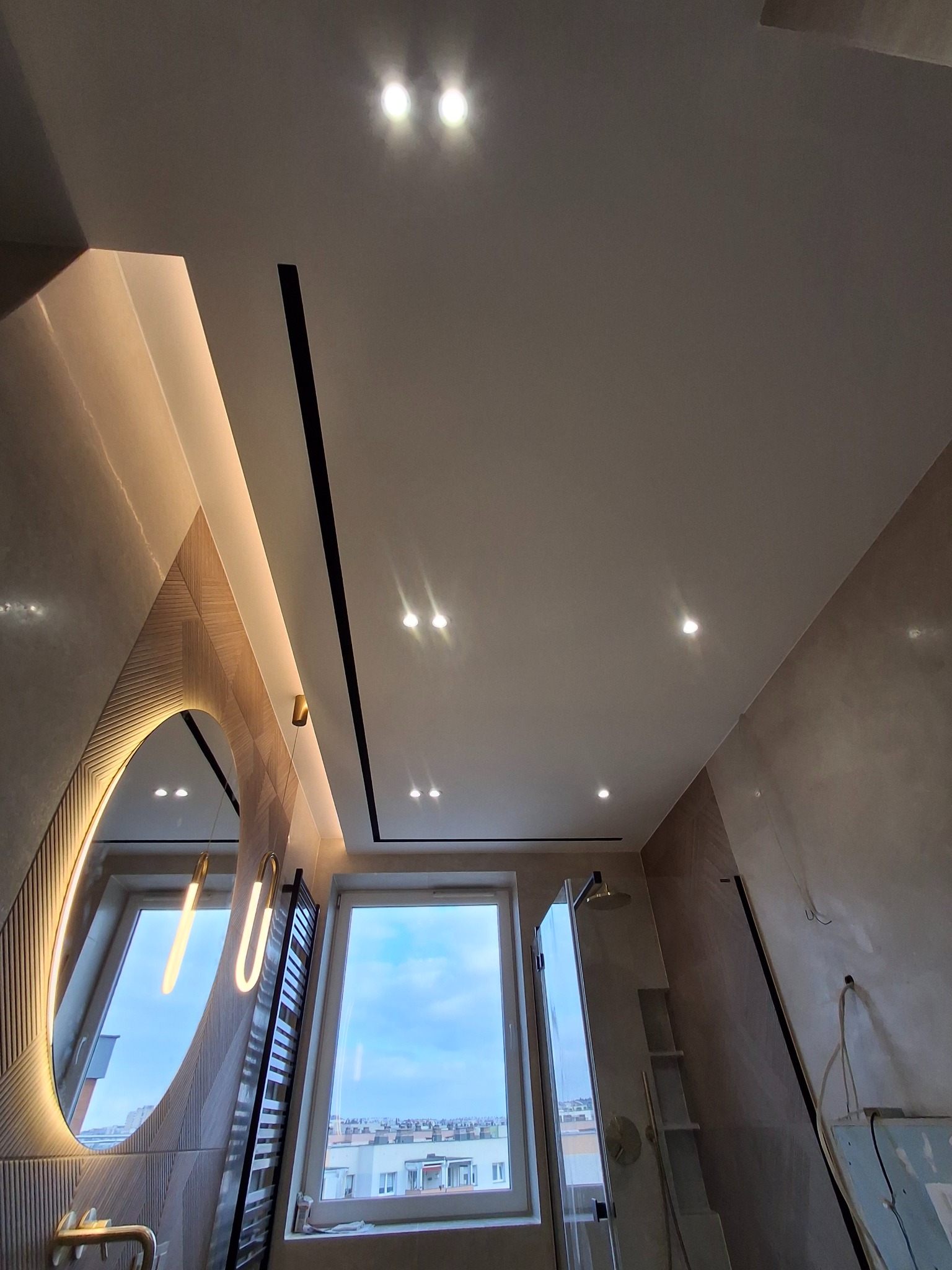 Nowoczesna łazienka w trakcie wykańczania: okrągłe lustro z podświetleniem LED, grzejnik drabinkowy, kabina prysznicowa ze złotą armaturą, okno z widokiem na miasto, sufit z oświetleniem punktowym,...