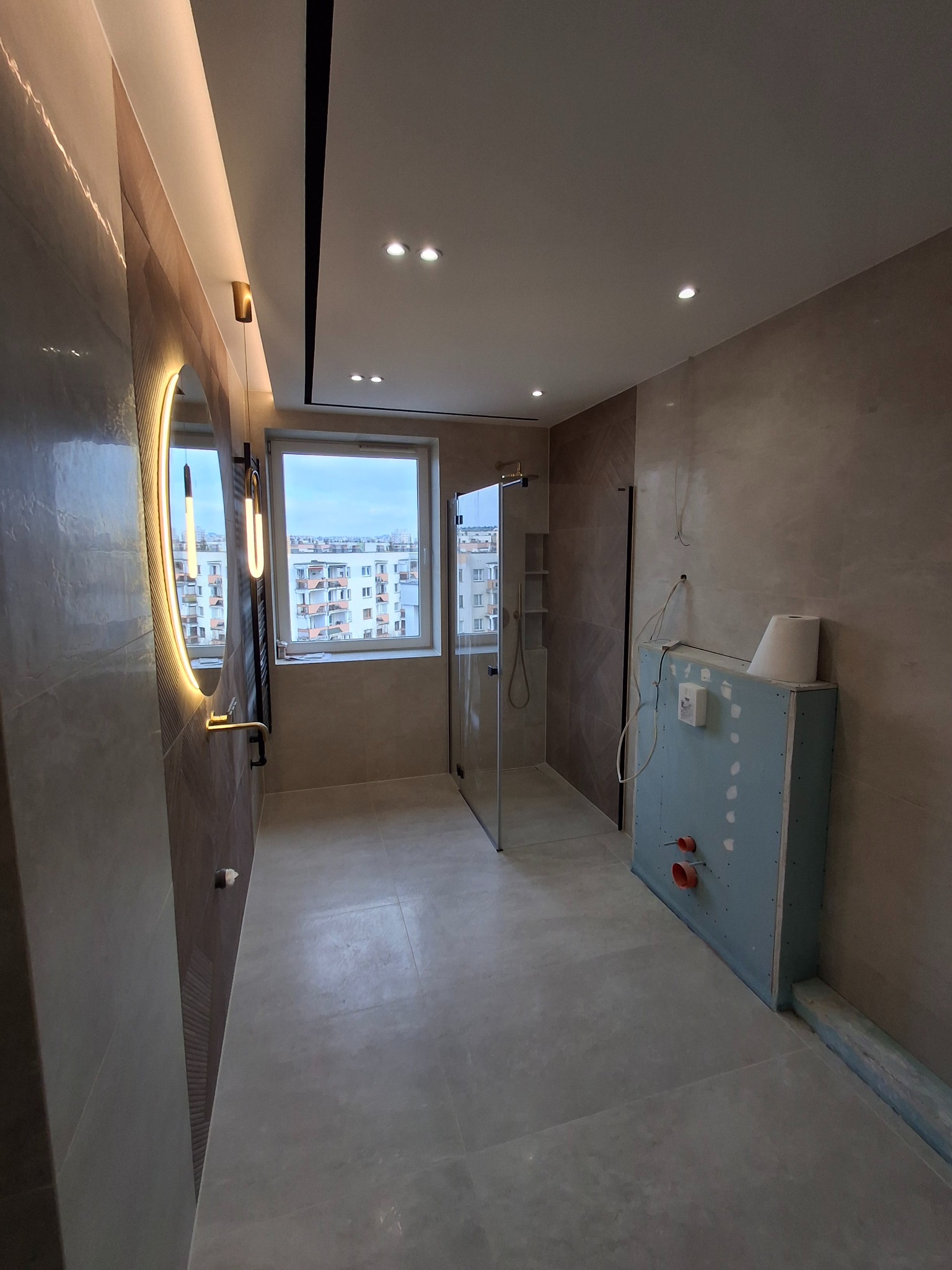 Wnętrze łazienki w trakcie wykańczania, widoczna kabina prysznicowa ze szklanymi drzwiami, okrągłe lustro z podświetleniem LED, zabudowa z płyty karton-gips z wystającymi rurami, beżowe płytki...