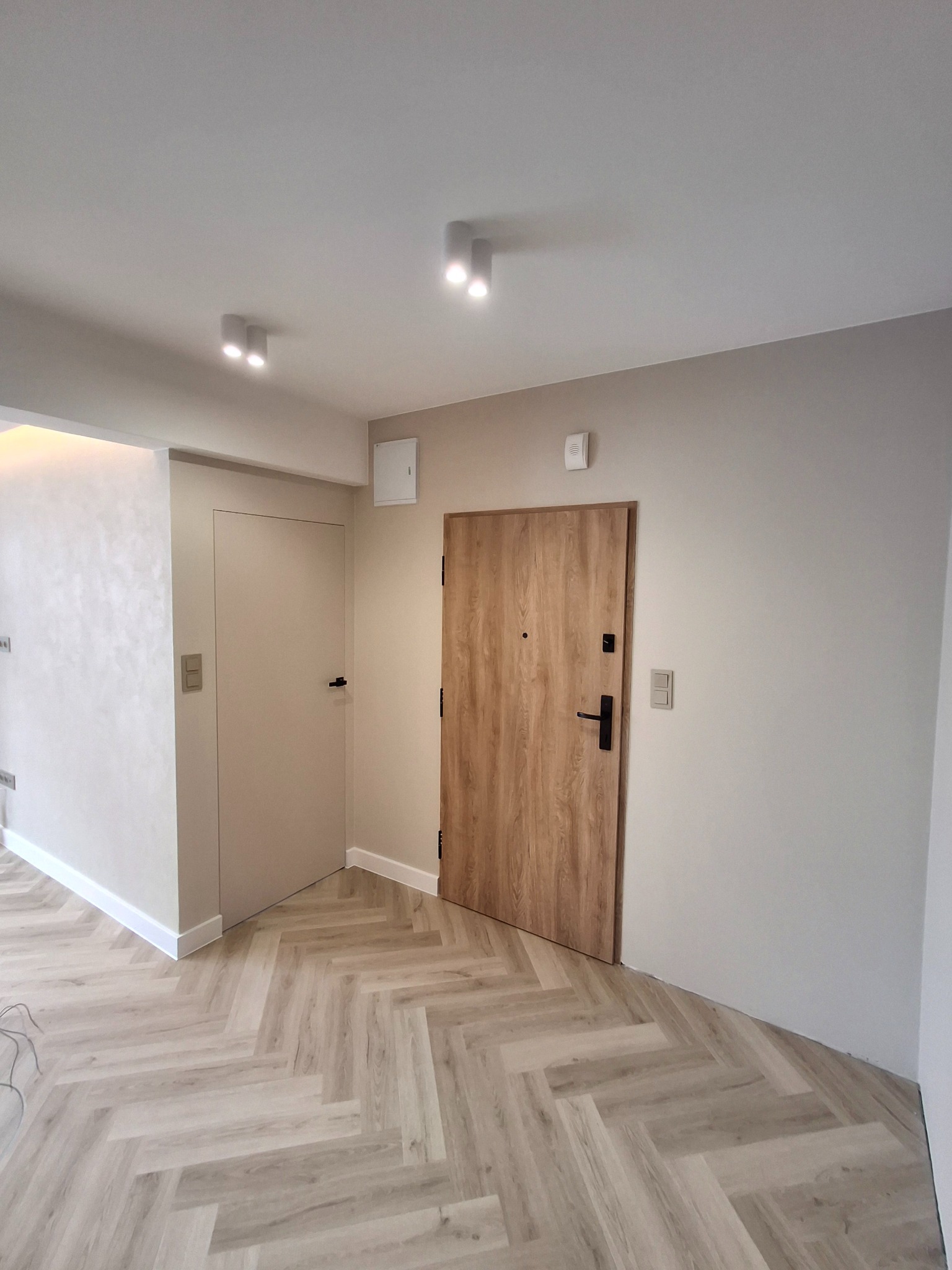 Beżowe wnętrze po odświeżeniu: parkiet ułożony w jodełkę, dwie pary punktowych lamp sufitowych i dwoje drzwi w odcieniach beżu.