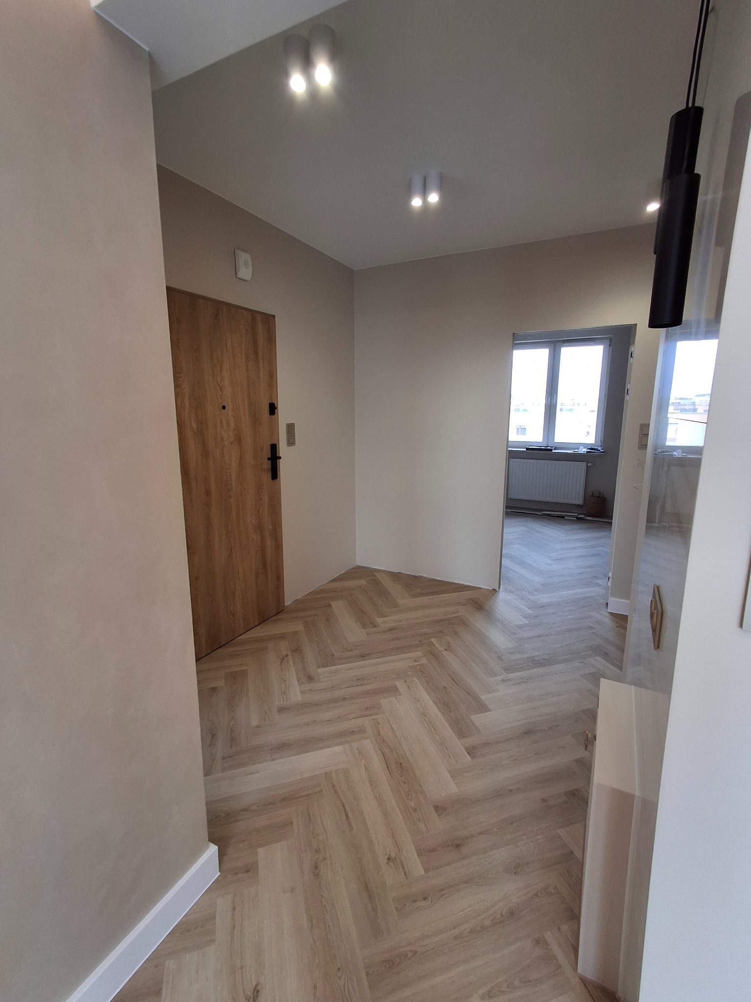 Wykończone wnętrze mieszkania w Kielcach z parkietem ułożonym w jodełkę, widoczny fragment ściany pomalowanej na beżowo, drewniane drzwi wejściowe oraz przejście do kolejnego pomieszczenia z oknem.