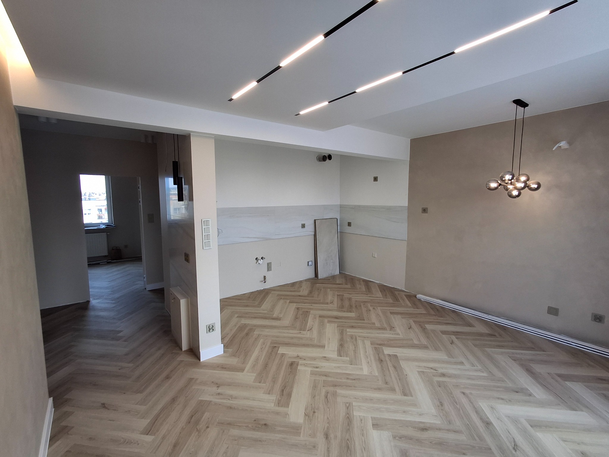 Jasne, przestronne wnętrze po remoncie z parkietem ułożonym w jodełkę, nowoczesnym oświetleniem sufitowym i częściowo wykończoną ścianą z płytkami.