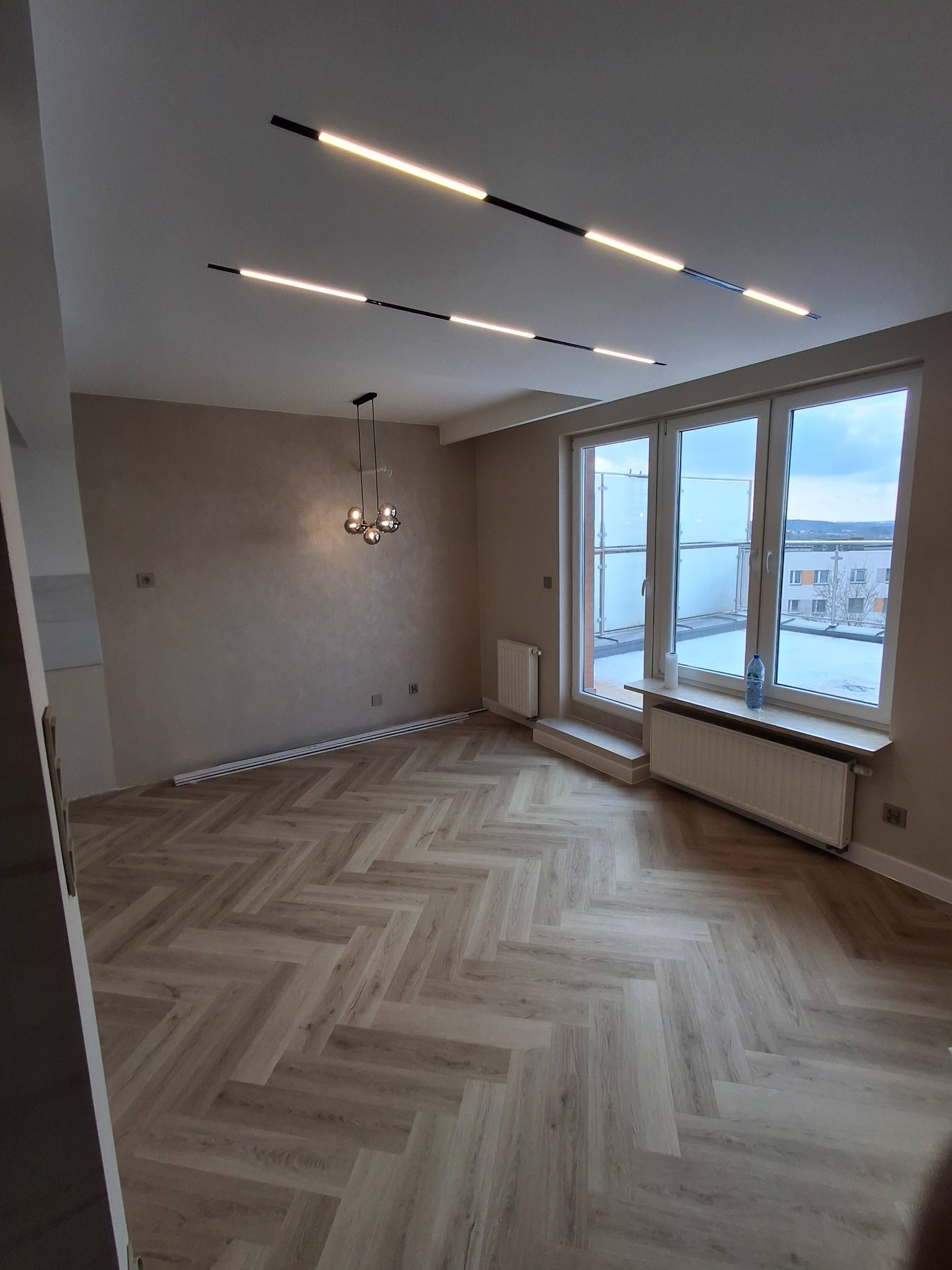 Nowoczesne wnętrze mieszkania po remoncie z parkietem ułożonym w jodełkę, minimalistycznym oświetleniem sufitowym i ścianami w odcieniach beżu. Widoczne grzejniki i okno balkonowe z widokiem...