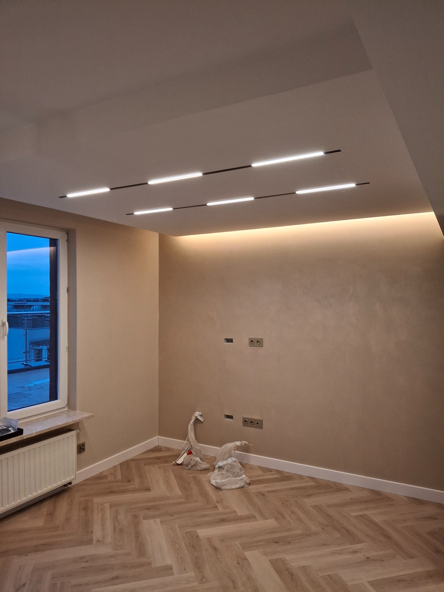 Wykończone wnętrze pokoju z parkietem w jodełkę, oświetleniem LED na suficie i ścianach w odcieniach beżu, widoczne gniazdka elektryczne i okno z widokiem na miasto w oddali.