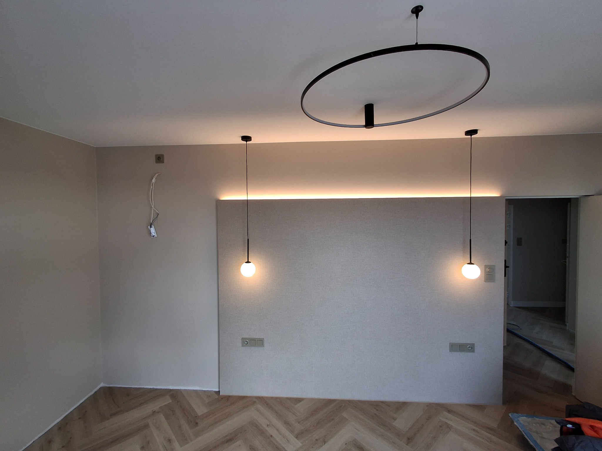 Wnętrze pokoju po remoncie z parkietem w jodełkę, oświetleniem LED za panelem ściennym i wiszącymi lampami kulami oraz okrągłą lampą sufitową.