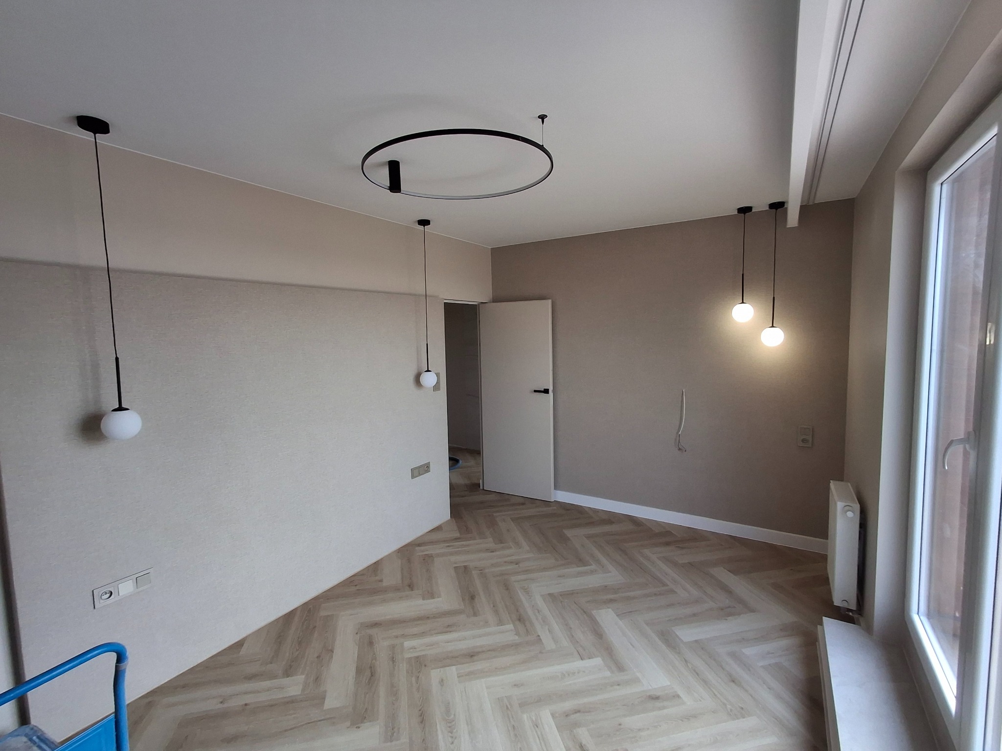 Wnętrze pokoju po remoncie z parkietem ułożonym w jodełkę, minimalistycznymi lampami wiszącymi i okrągłą lampą sufitową, widoczne gniazdka elektryczne i grzejnik.