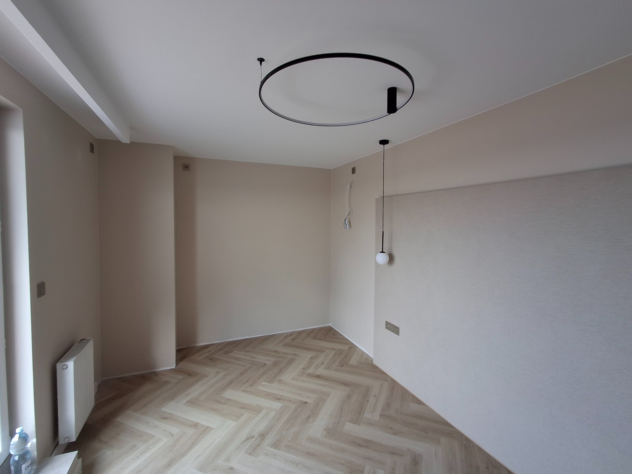 Jasne, puste pomieszczenie po remoncie z podłogą ułożoną w jodełkę, ścianami w odcieniach beżu, nowoczesnym okrągłym oświetleniem sufitowym i grzejnikiem przy oknie z butelką wody mineralnej obok.