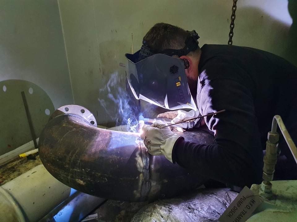 Pracownik w masce spawalniczej łączy rurę grubościenną metodą spawania łukowego, widoczne iskry i dym, wnętrze pomieszczenia technicznego.