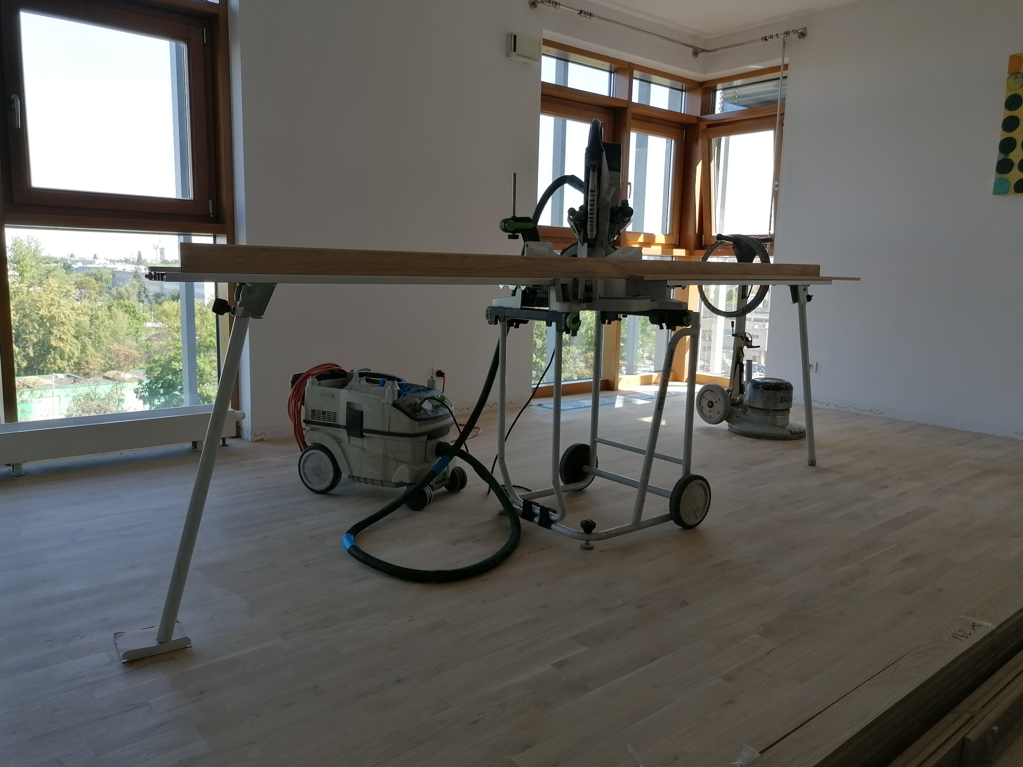 Układanie jasnego parkietu z użyciem frezarki na stelażu, odkurzacza przemysłowego i cykliniarki w jasnym, przestronnym wnętrzu z dużymi oknami.