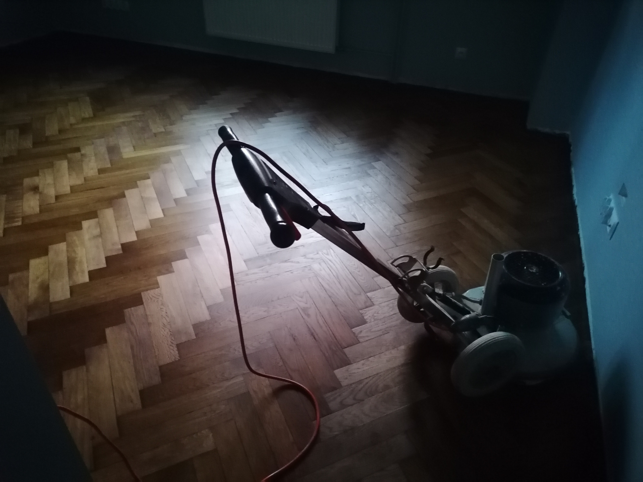 Parkiet ułożony w jodełkę, w trakcie cyklinowania, widoczna maszyna do cyklinowania z czerwonym przewodem, oświetlona punktowo.