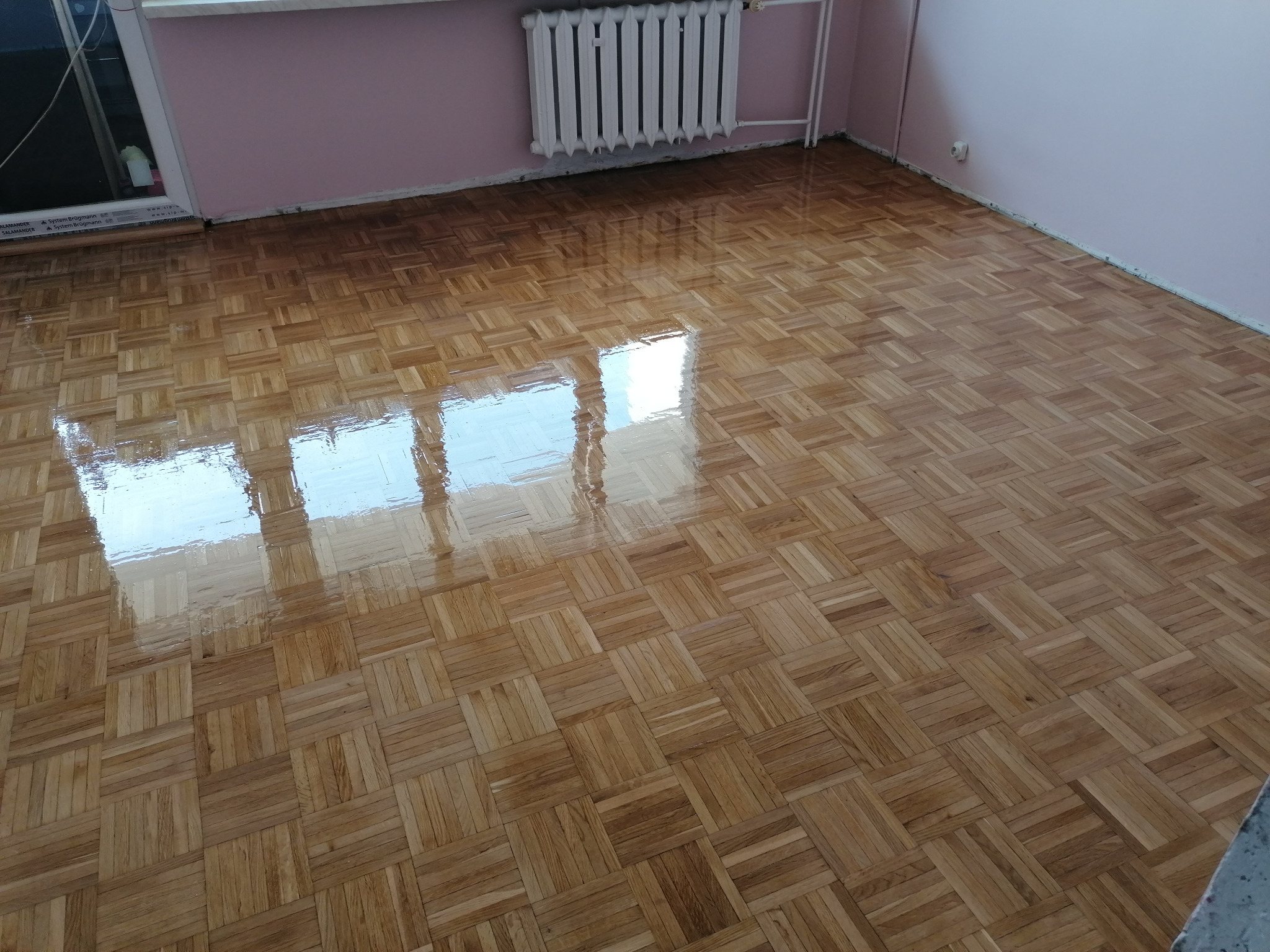 Świeżo polakierowana podłoga z parkietu ułożonego w kwadraty, widoczne odbicie okna, różowe ściany i biały grzejnik.