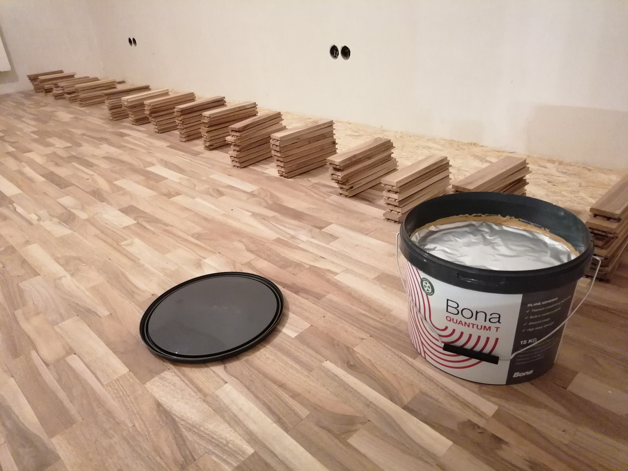 Układanie parkietu z klejem Bona Quantum T w wiadrze, widoczne ułożone deski parkietowe i czarna pokrywa od wiadra na podłodze.
