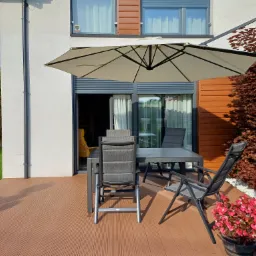 Zadaszony taras z kompozytowej deski, szare meble ogrodowe (stół i krzesła), duży parasol przeciwsłoneczny, ozdobny klon palmowy w tle, donica z czerwonymi kwiatami.