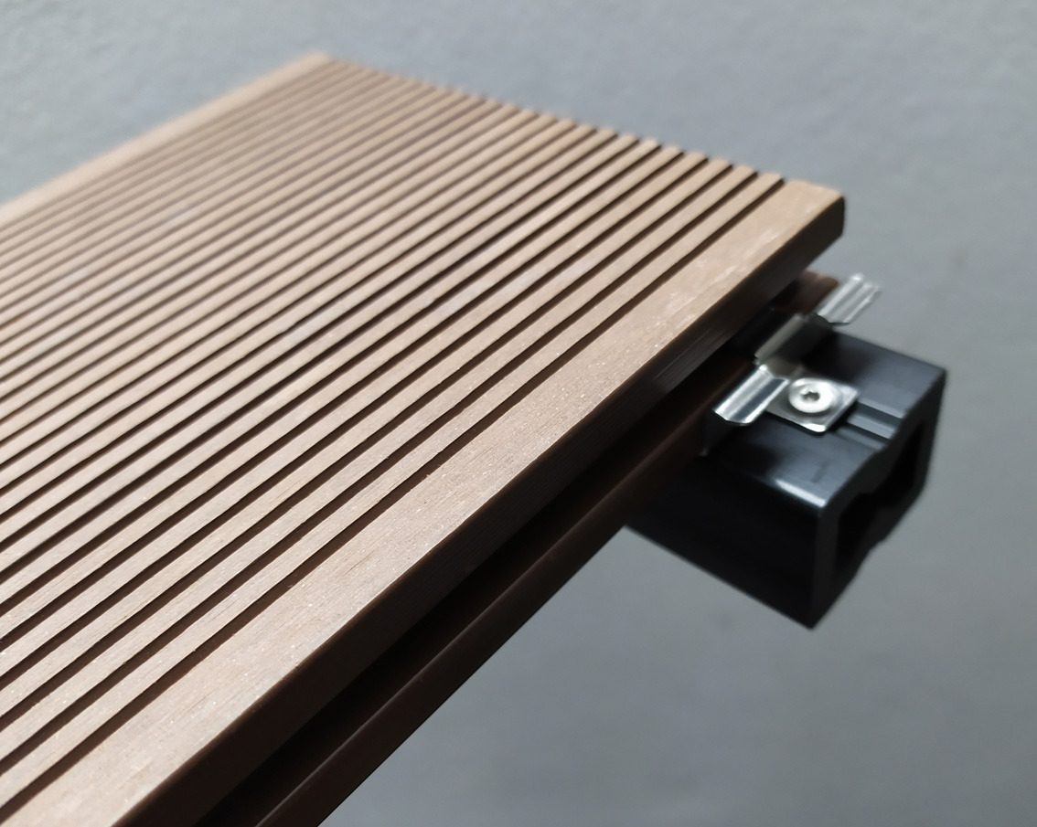 Detale montażu deski kompozytowej tarasowej: widoczne ryflowanie, czarny klips montażowy z metalowym uchwytem i śrubą.