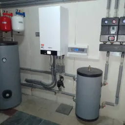 Biały piec grzewczy Viessmann zamontowany na ścianie, obok dwa szare zbiorniki na wodę, czerwony i biały zbiornik wyrównawczy oraz szara instalacja rurowa z czujnikami i zaworami w kotłowni.