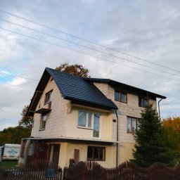 USŁUGI DEKARSKIE PIOTR BĄBEL - Dwupiętrowy dom w trakcie budowy z nowym, czarnym dachem z blachodachówki, widoczne bloczki betonowe i brak elewacji, drewniane okna, ogrodzenie z brązowych sztachet i przyczepa kempingowa w tle.