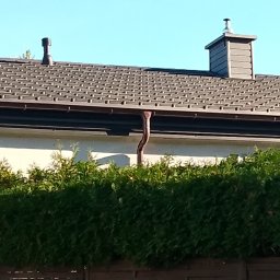 USŁUGI DEKARSKIE PIOTR BĄBEL - Brązowy dach z blachodachówki z dwoma kominami i rynną miedzianą, widoczny zza żywopłotu.