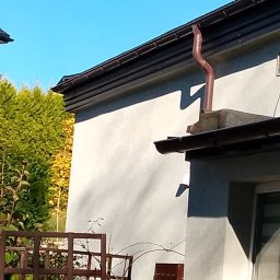 USŁUGI DEKARSKIE PIOTR BĄBEL - Brązowa rynna spustowa zamontowana na dachu budynku jednorodzinnego, widoczna na tle błękitnego nieba i zielonych drzew.