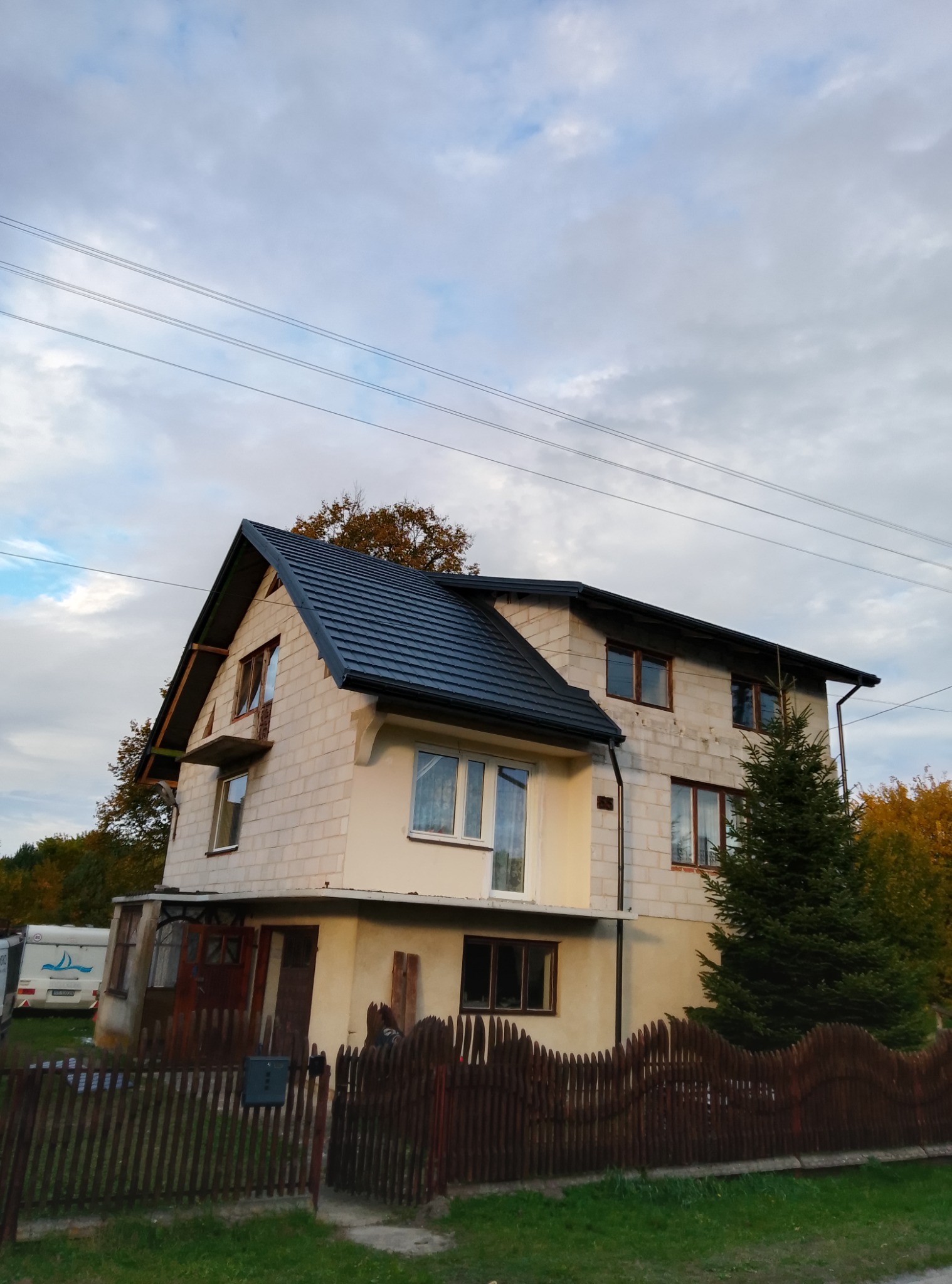 Dwupiętrowy dom w trakcie budowy z nowym, czarnym dachem z blachodachówki, widoczne bloczki betonowe i brak elewacji, drewniane okna, ogrodzenie z brązowych sztachet i przyczepa kempingowa w tle.