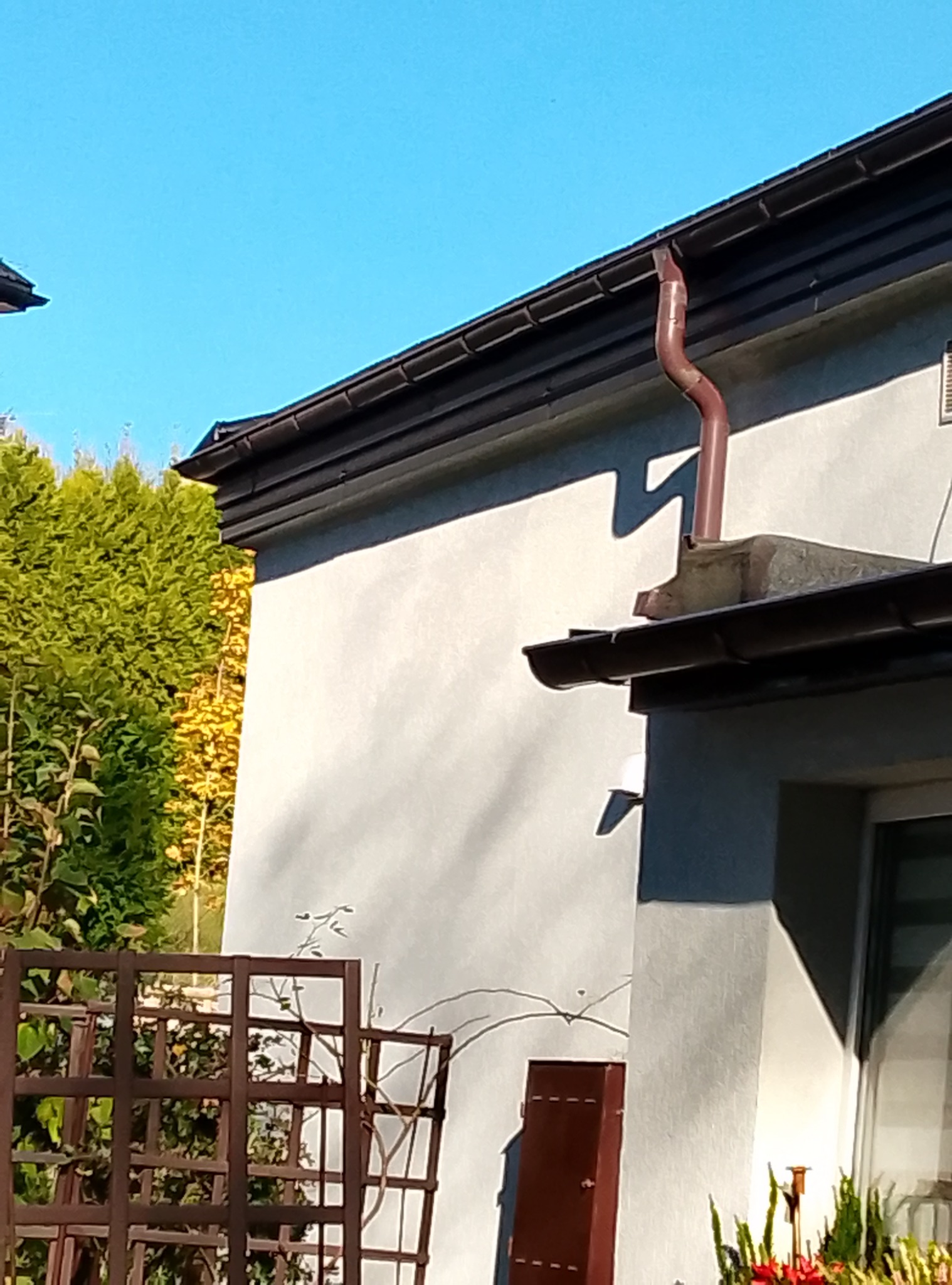 Brązowa rynna spustowa zamontowana na dachu budynku jednorodzinnego, widoczna na tle błękitnego nieba i zielonych drzew.