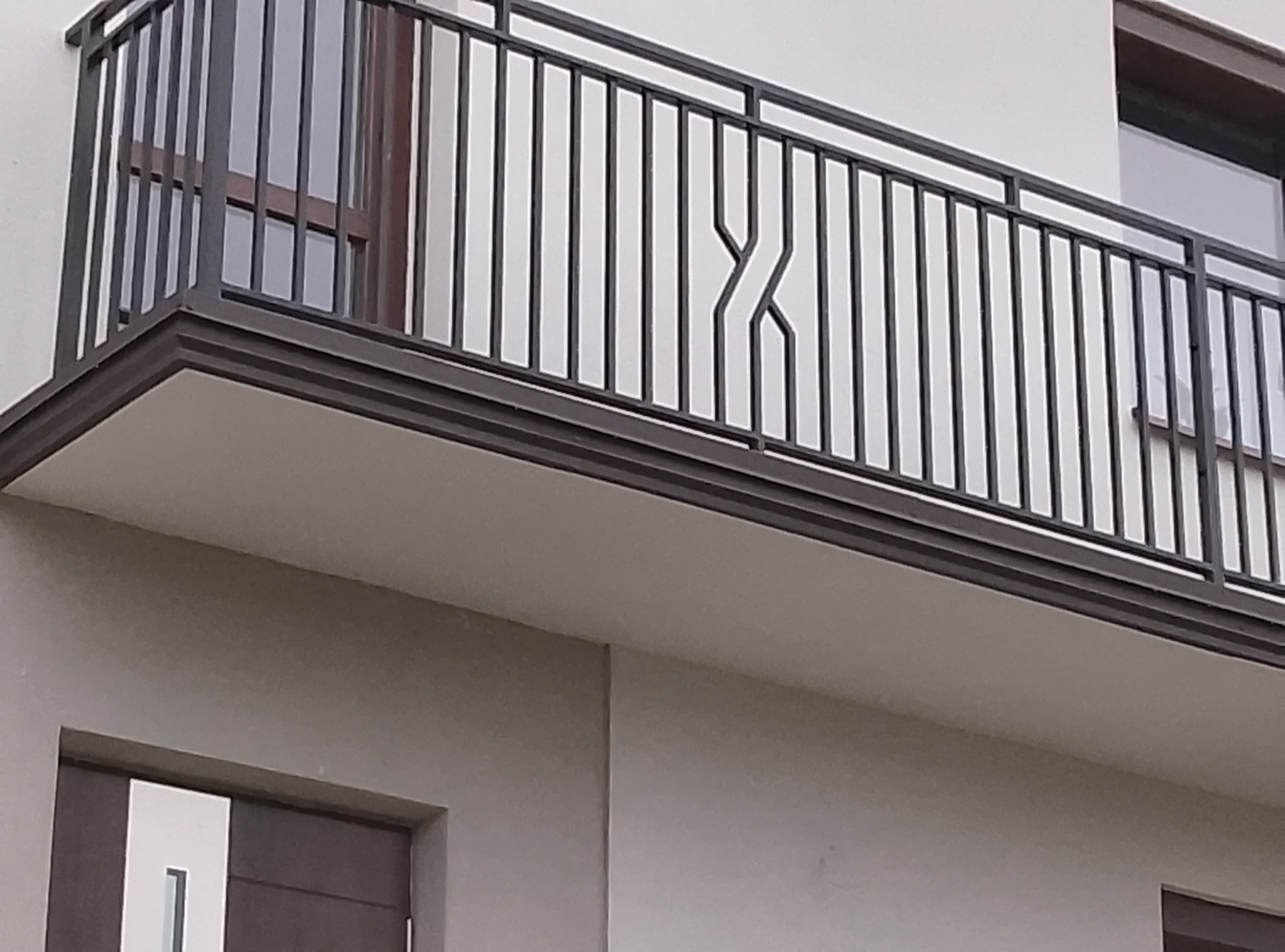 Nowoczesna balustrada balkonowa z czarnych, pionowych prętów z ozdobnym motywem geometrycznym na tle białej elewacji budynku.