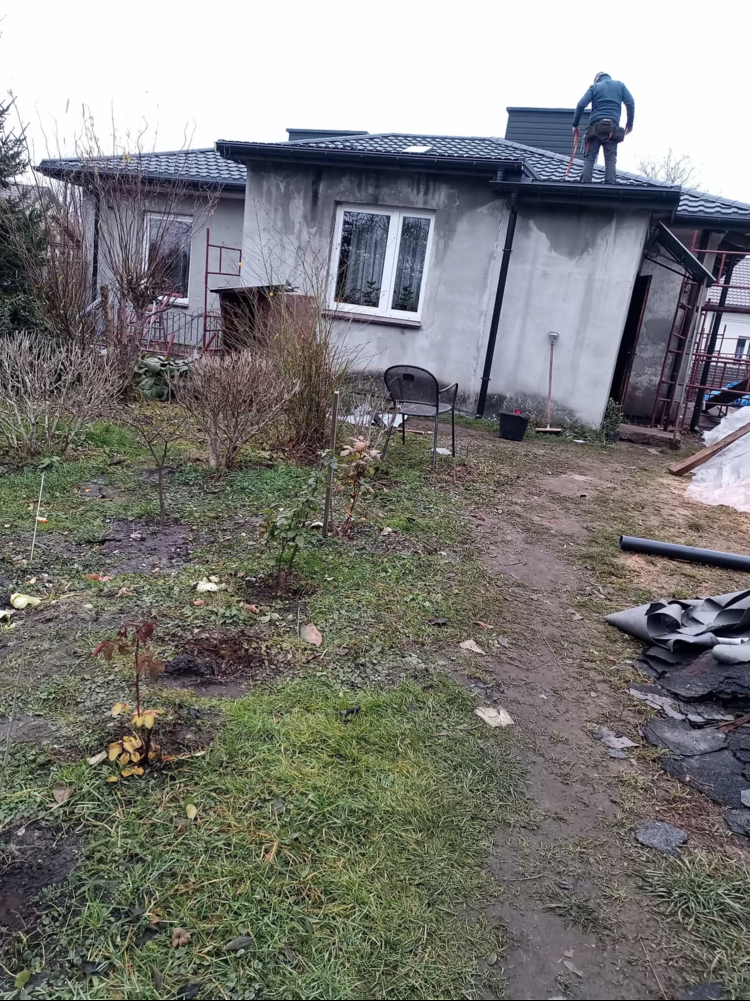 Dekarz na dachu domu jednorodzinnego w trakcie montażu rynny, widoczne narzędzia i fragmenty nowego pokrycia dachowego, teren wokół domu zaniedbany, pochmurny dzień.