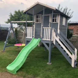 Drewniany plac zabaw dla dzieci w ogrodzie, z domkiem na podeście, zjeżdżalnią i huśtawką, pomalowany na szaro z białymi detalami, na tle trawnika i drzew.