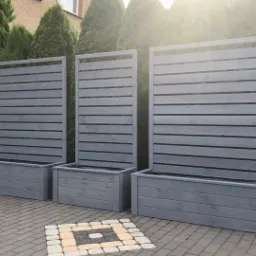 Trzy szare, drewniane donice z panelami ażurowymi, ustawione na kostce brukowej, z iglakami w tle, tworzące element dekoracyjny ogrodu.