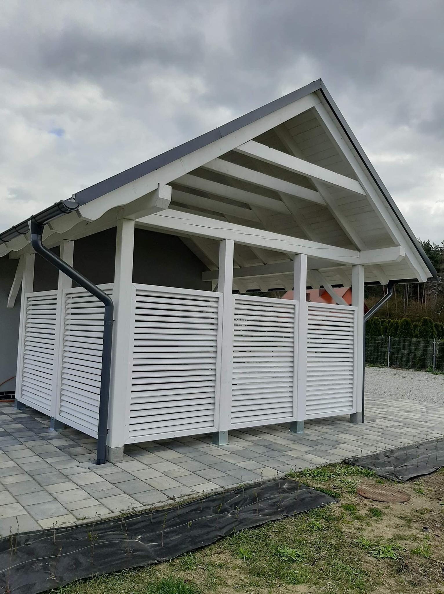 Biała konstrukcja z drewnianymi lamelami i szarym dachem, prawdopodobnie wiata lub altana, na kostce brukowej, widok z zewnątrz w pochmurny dzień.