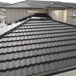 Nowy, błyszczący dach z metalowej dachówki w kolorze grafitowym, widok z góry, w tle dom w trakcie budowy z widocznymi rusztowaniami i izolacją przeciwwiatrową.