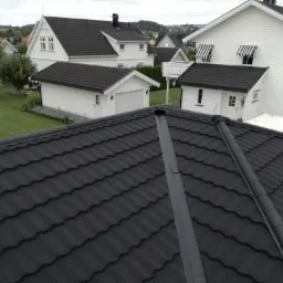 Widok z góry na nowo położony, czarny dach z blachodachówki modułowej, w tle białe domy jednorodzinne z ciemnymi dachami i zielony ogród.