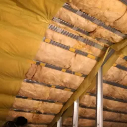 Montaż izolacji z wełny mineralnej w skośnym dachu pod żółtą folią paroizolacyjną, widoczne metalowe profile konstrukcyjne i pracownik w czarnej bluzie.