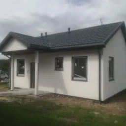 Niski, parterowy budynek z jasną elewacją i ciemnoszarą dachówką, widoczne okna z folią ochronną, teren wokół w trakcie zagospodarowania.