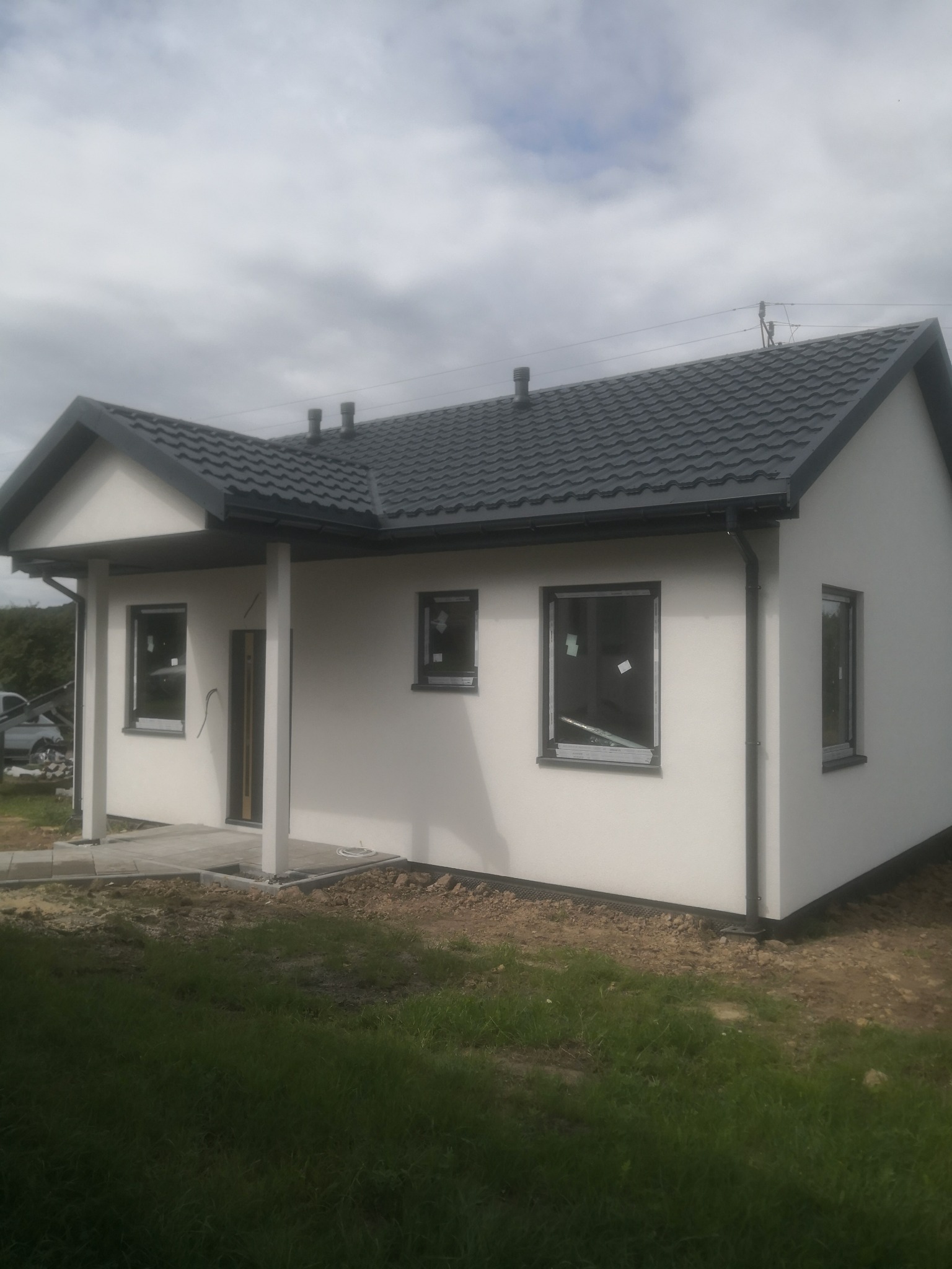 Niski, parterowy budynek z jasną elewacją i ciemnoszarą dachówką, widoczne okna z folią ochronną, teren wokół w trakcie zagospodarowania.