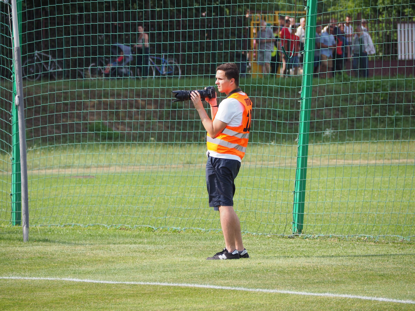 Młody mężczyzna w pomarańczowej kamizelce z napisem 'FOTO' trzymający duży aparat fotograficzny na tle siatki ogrodzeniowej i grupy osób w oddali.