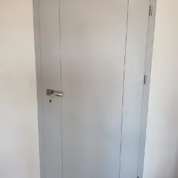 Zamknięte, szare drzwi wewnętrzne z prostokątnymi panelami, zamontowane w białej ścianie, widoczny srebrny klamek i zawiasy.