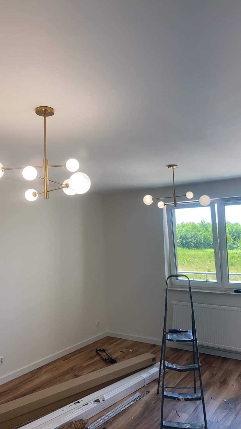 Nowoczesne złote lampy sufitowe z białymi kulami w pokoju z panelami podłogowymi, drabina i materiały budowlane przy ścianie.