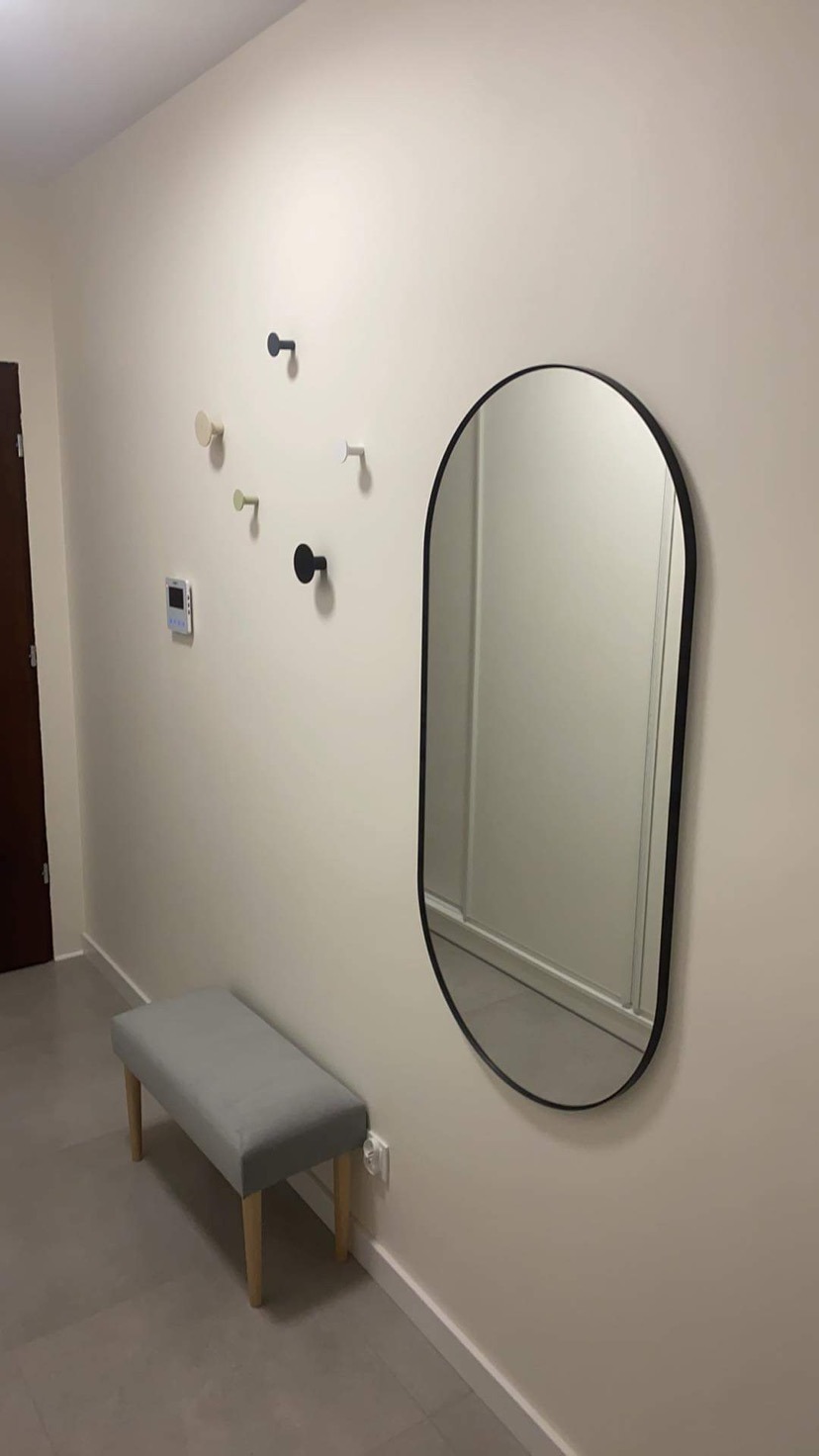 Wnętrze przedpokoju z kremowymi ścianami, owalnym lustrem w czarnej ramie, szarą tapicerowaną ławką, oraz zestawem wieszaków w różnych kolorach, tworzących dekoracyjny układ na ścianie.