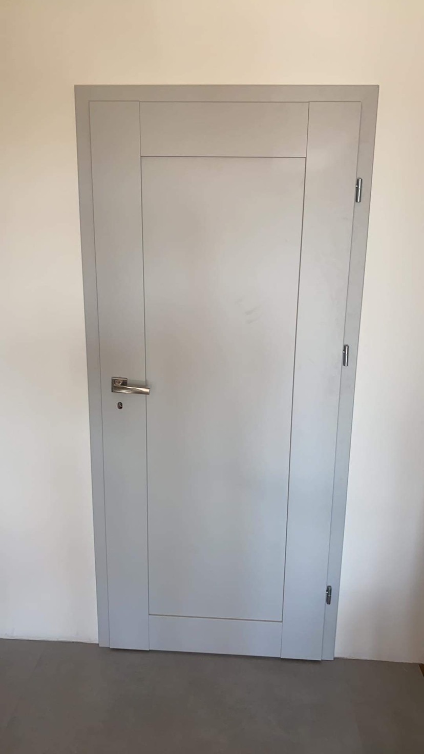 Zamknięte, szare drzwi wewnętrzne z prostokątnymi panelami, zamontowane w białej ścianie, widoczny srebrny klamek i zawiasy.