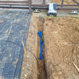 Wykop w ziemi wzdłuż bramy przesuwnej z widocznym kablem w niebieskiej rurze ochronnej, przygotowanie do podłączenia napędu bramy.